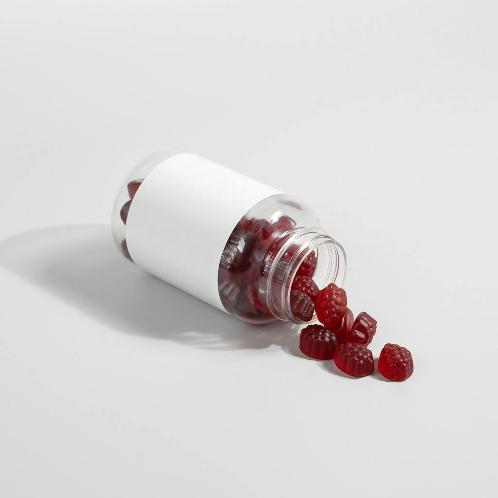 A transparent bottle with red gummy supplements spilling onto a white surface.